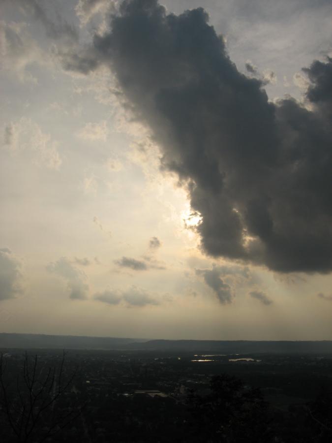 A moody sky with a large dark cloud obscuring the sun, casting a soft light over a landscape below. A distant horizon is visible with patches of land and water, surrounded by a hazy atmosphere. The scene captures a tranquil yet dramatic moment in nature. Bluff Trails mountain bike trail.