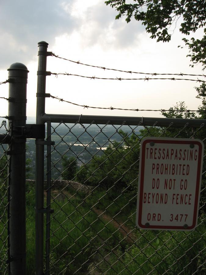 A chain-link fence topped with barbed wire, enclosing a view of a landscape below. A sign on the fence reads, "TRESPASSING PROHIBITED DO NOT GO BEYOND FENCE ORD. 3477." The sky is partly cloudy, and greenery is visible in the foreground. Bluff Trails mountain bike trail.