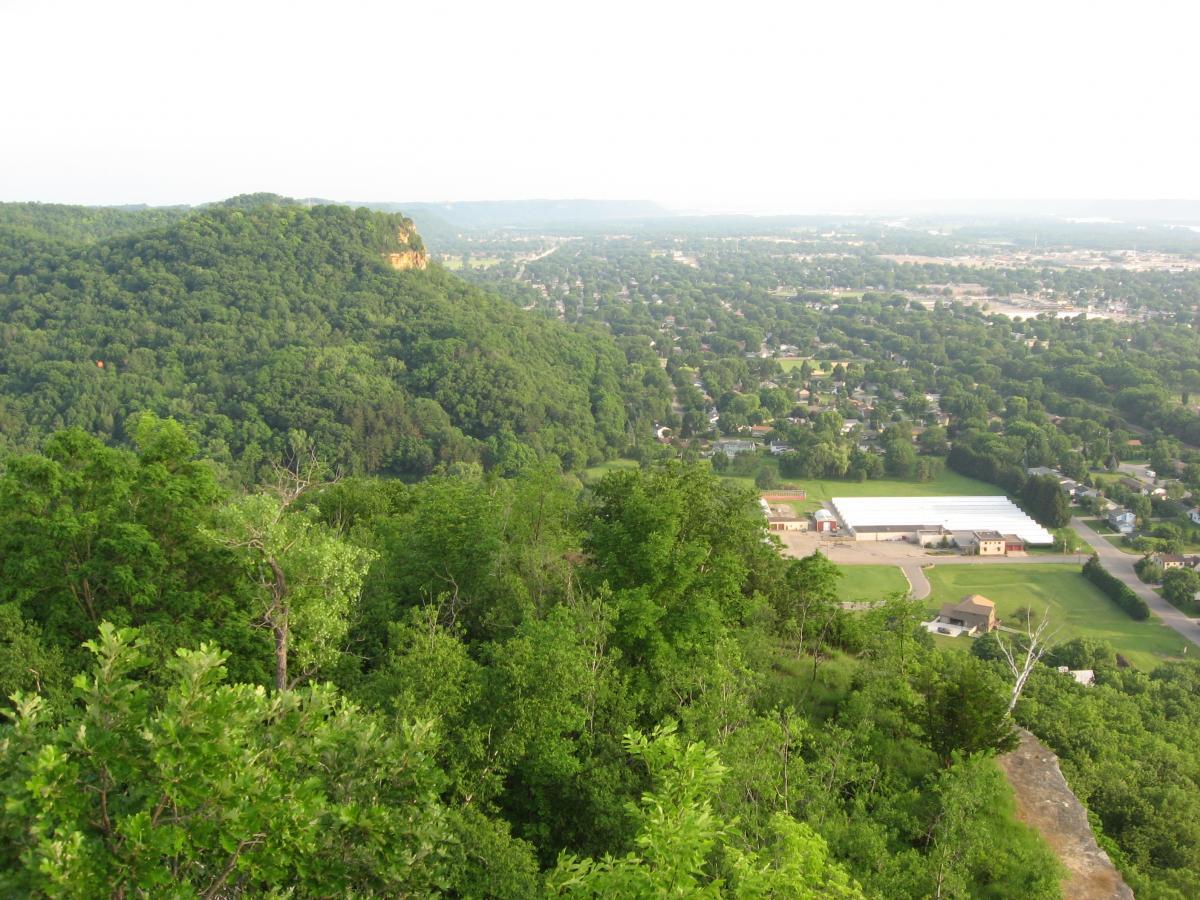 A scenic view from a high vantage point overlooking lush green hills and a valley. In the foreground, dense trees and foliage are visible, while in the background, a small town can be seen with houses, a sports field, and open fields. The horizon features rolling hills and a clear sky. Bluff Trails mountain bike trail.