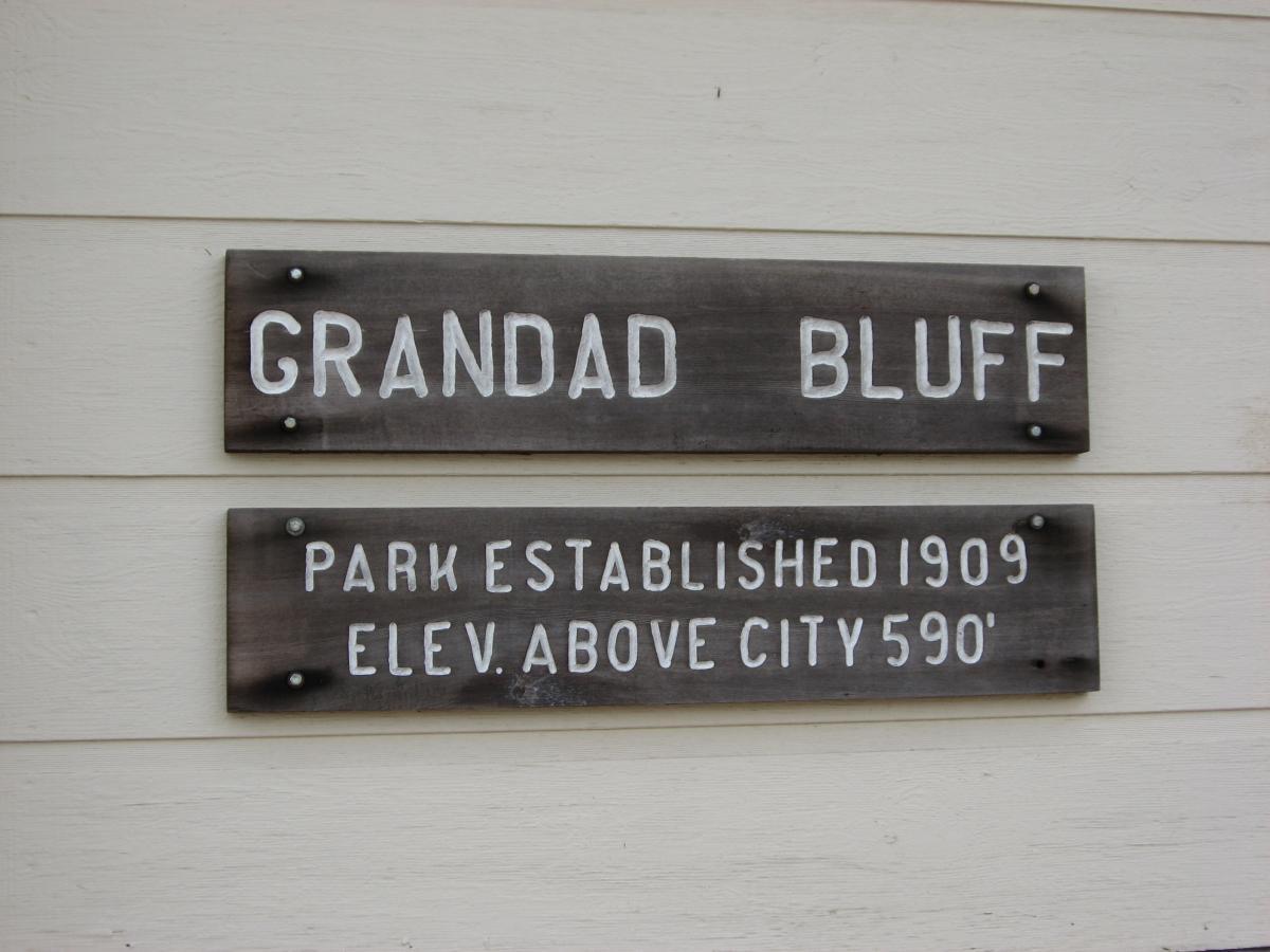 Two wooden signs mounted on a wall. The upper sign reads "GRANDAD BLUFF" in large, white letters. The lower sign states "PARK ESTABLISHED 1909" and "ELEV. ABOVE CITY 590'" in smaller white letters. Bluff Trails mountain bike trail.