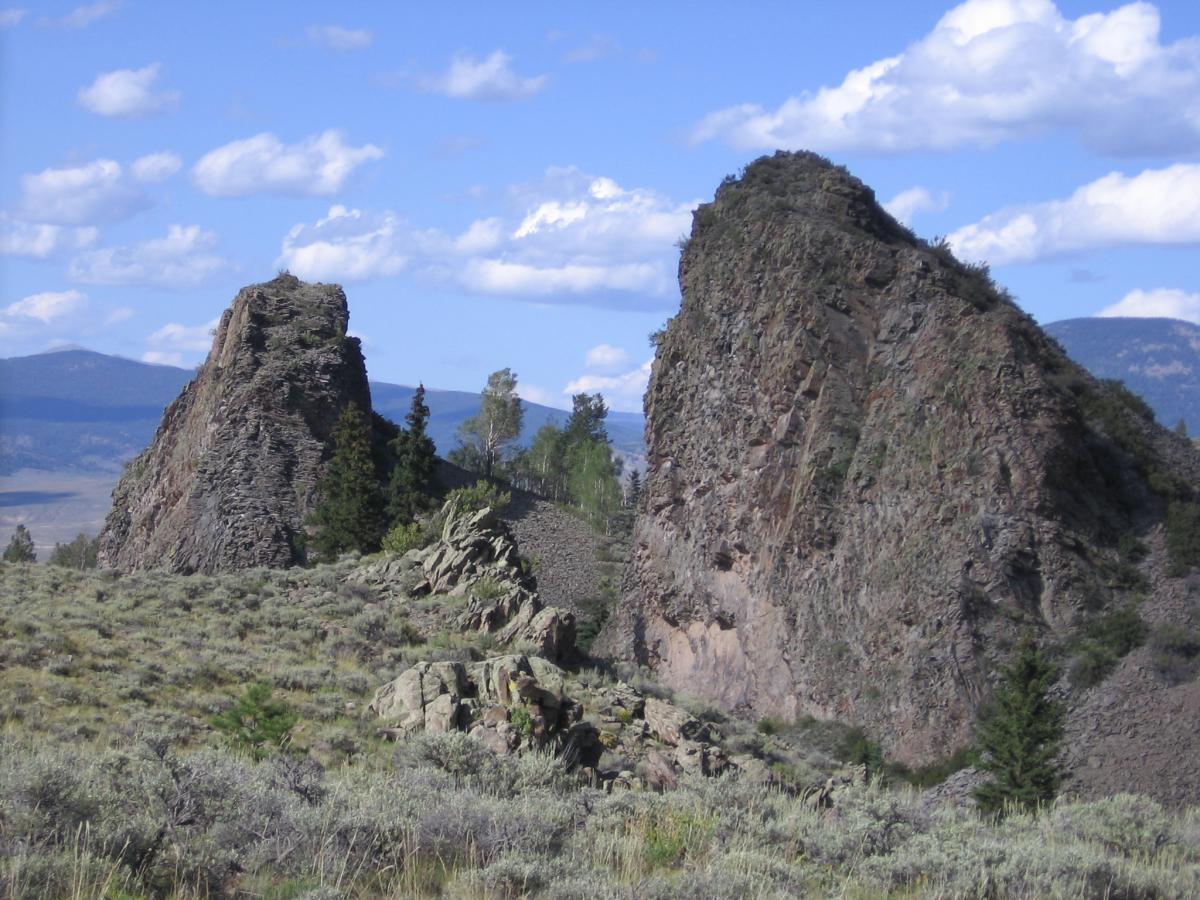 Two prominent rock formations rise above a grassy landscape, surrounded by sparse trees and under a partly cloudy blue sky. The formations exhibit jagged edges and rugged textures, set against distant mountains in the background. Right Hand Creek mountain bike trail.