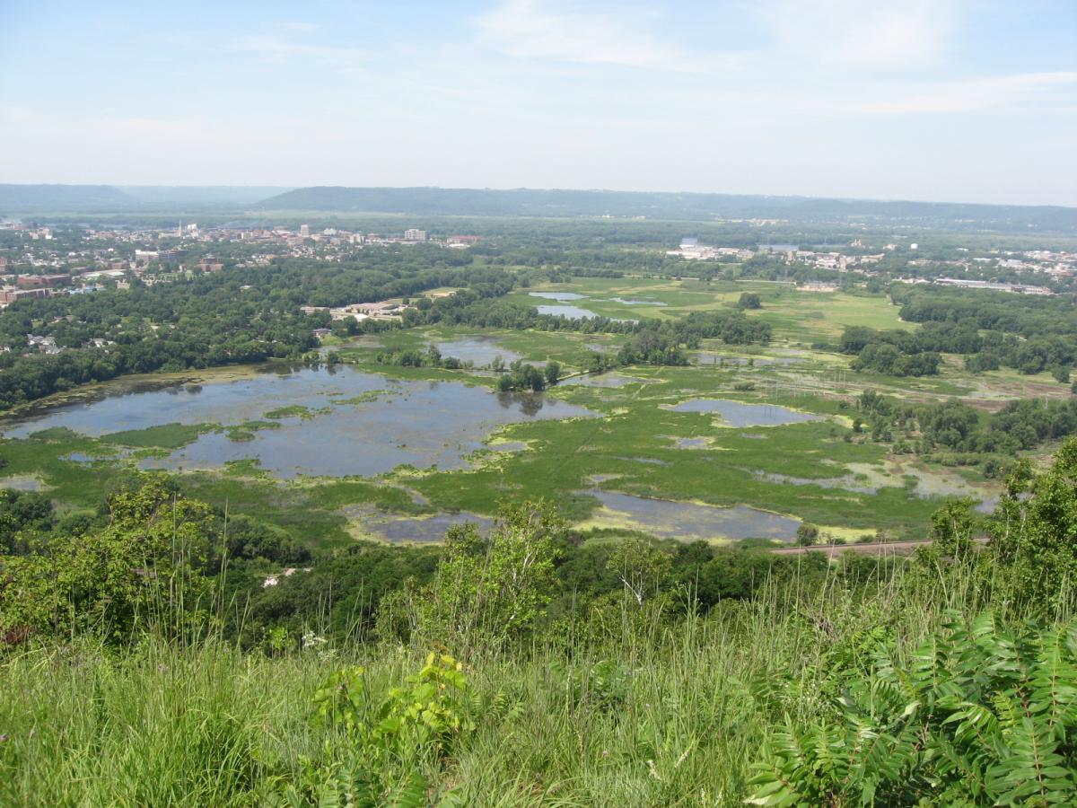 A panoramic view of a lush green landscape featuring several ponds surrounded by wetlands. In the background, a small town is visible nestled among the trees, with rolling hills beyond. The sky is bright blue with scattered clouds, creating a picturesque natural scene. Bluff Trails mountain bike trail.
