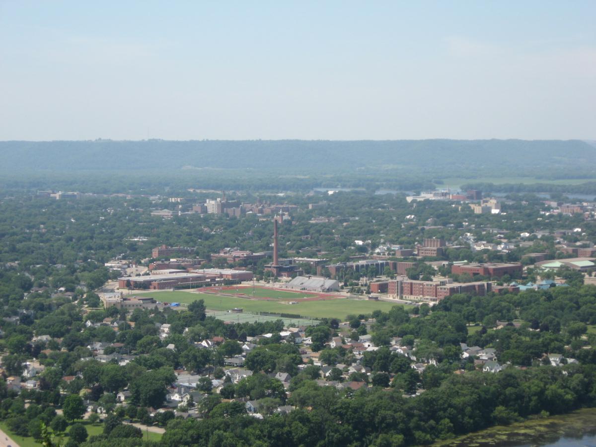 Aerial view of a green landscape showing a city with a mix of urban structures and residential areas. In the foreground, there is a sports field surrounded by buildings, with a distinct smokestack visible. The background features rolling hills and a clear blue sky, suggesting a sunny day. Bluff Trails mountain bike trail.