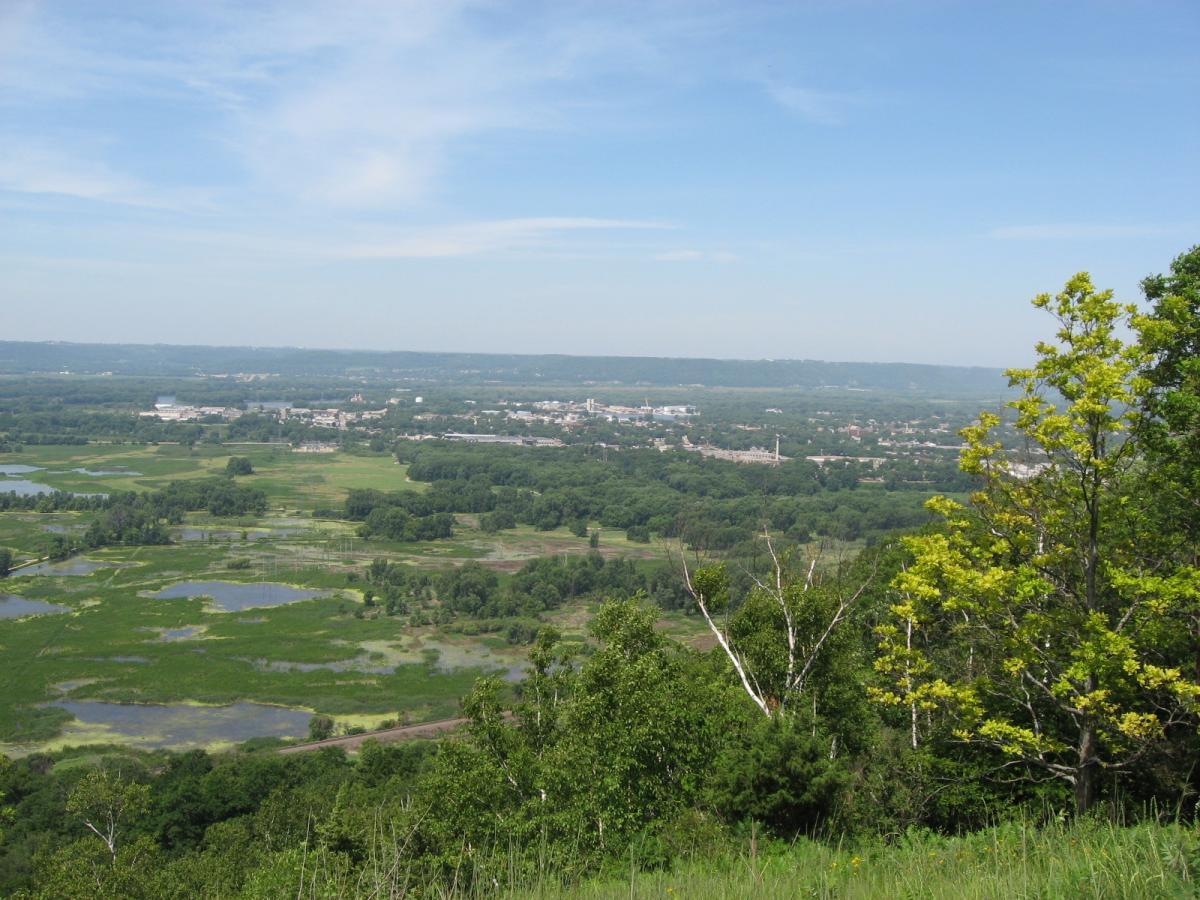A panoramic view from a hill overlooking a lush, green landscape with wetlands and patches of water. In the distance, a small town is visible under a clear blue sky with a few wispy clouds. Trees in the foreground provide a natural frame to the scene. Bluff Trails mountain bike trail.