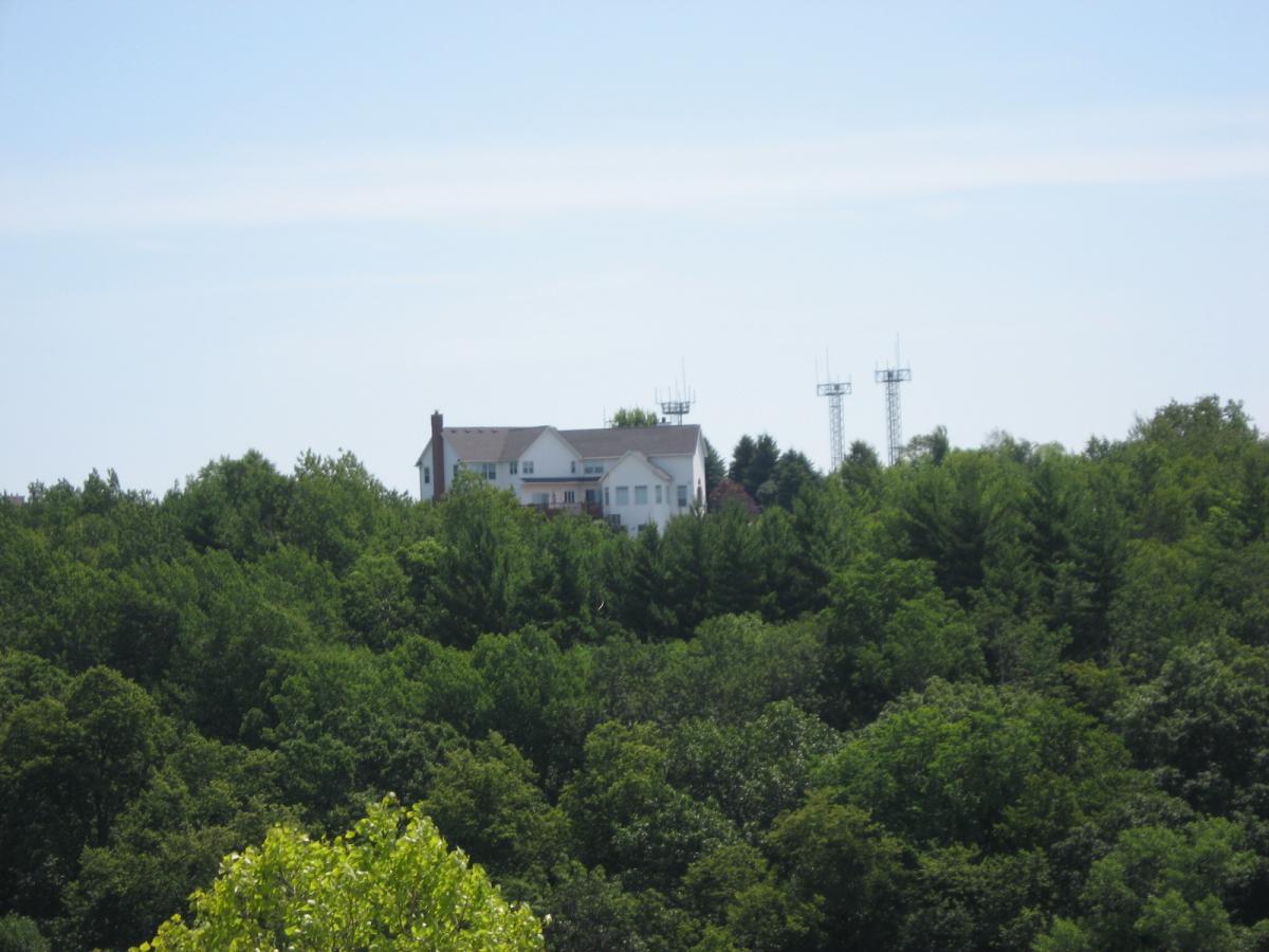 A large house situated atop a hill, surrounded by lush green trees and vegetation, with two communication towers visible nearby against a clear blue sky. Bluff Trails mountain bike trail.
