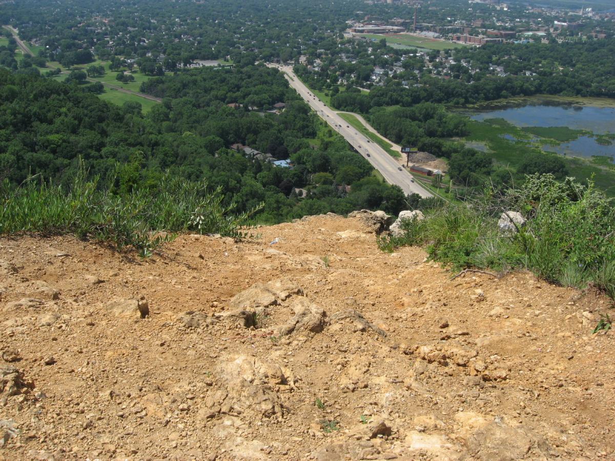 Alt text: A view from the edge of a rocky hillside, showing a landscape of trees and a winding road below. In the distance, a small town is visible with a mix of residential homes and open spaces. The clear sky suggests a sunny day. Bluff Trails mountain bike trail.