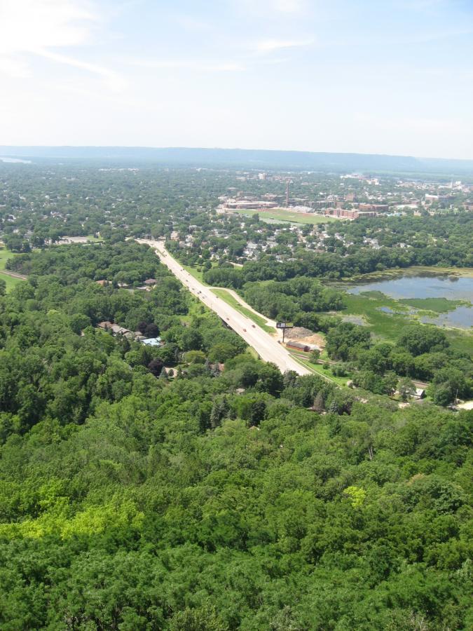 A panoramic view of a green landscape with dense trees, a winding road, and a small town in the distance. A lake can be seen to the right, with buildings and structures visible in the background under a clear blue sky. Bluff Trails mountain bike trail.