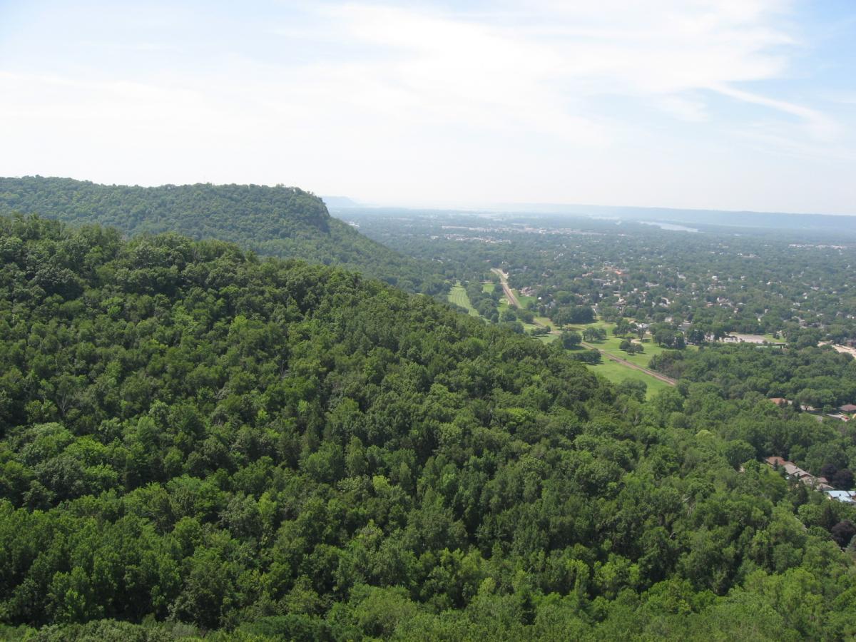 A panoramic view of a lush green landscape featuring rolling hills and dense forests, with a residential area visible in the distance. The sky is bright with a few clouds, and a river can be seen meandering through the valley. Bluff Trails mountain bike trail.