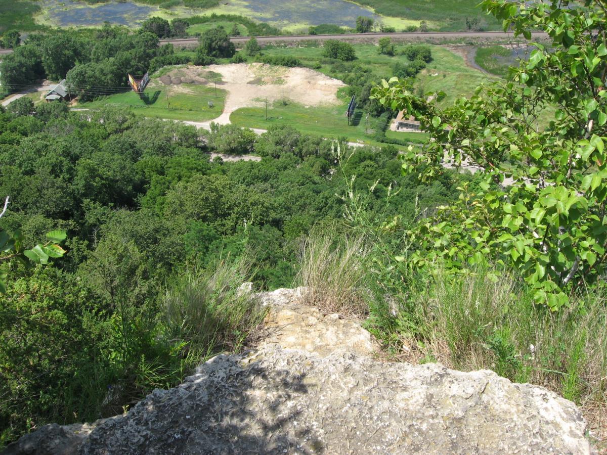 A scenic view from the edge of a rocky outcrop overlooking a lush green landscape, featuring trees and open fields. In the distance, a dirt area and a path are visible, surrounded by greenery. The sky is clear and bright, suggesting a sunny day. Bluff Trails mountain bike trail.