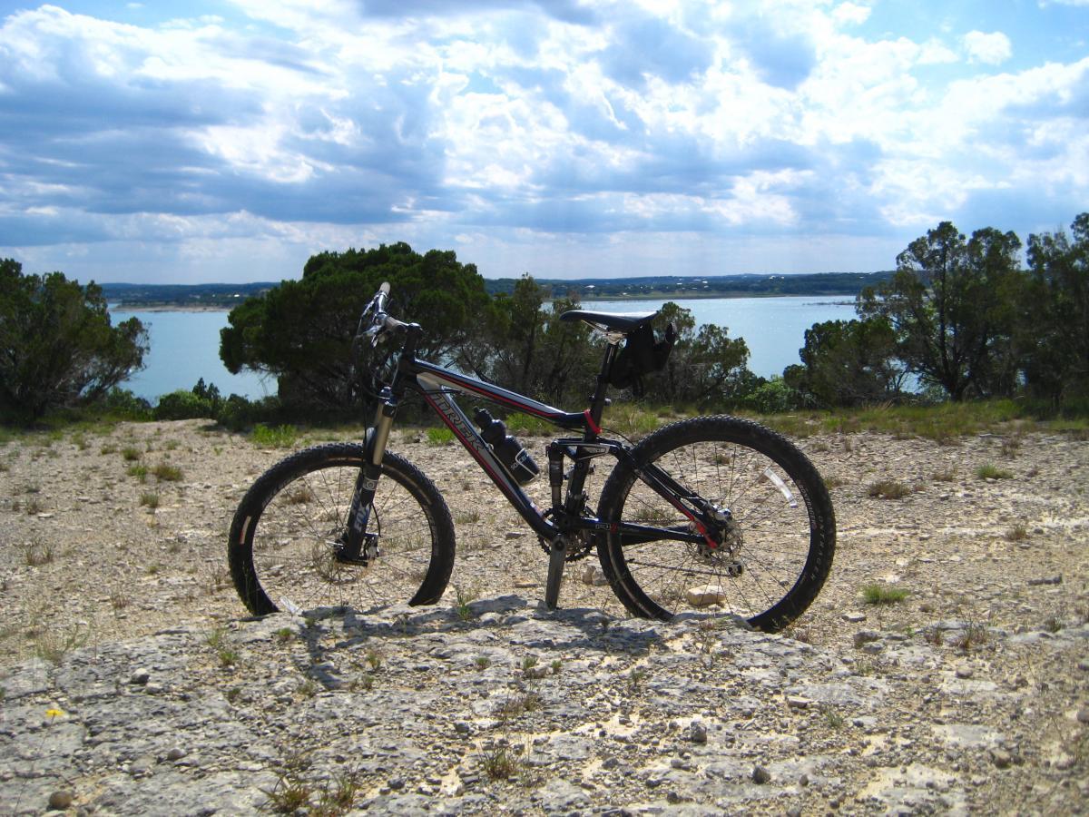 A mountain bike resting on rocky terrain with a lake and trees in the background under a cloudy sky. Madrone Trail mountain bike trail.