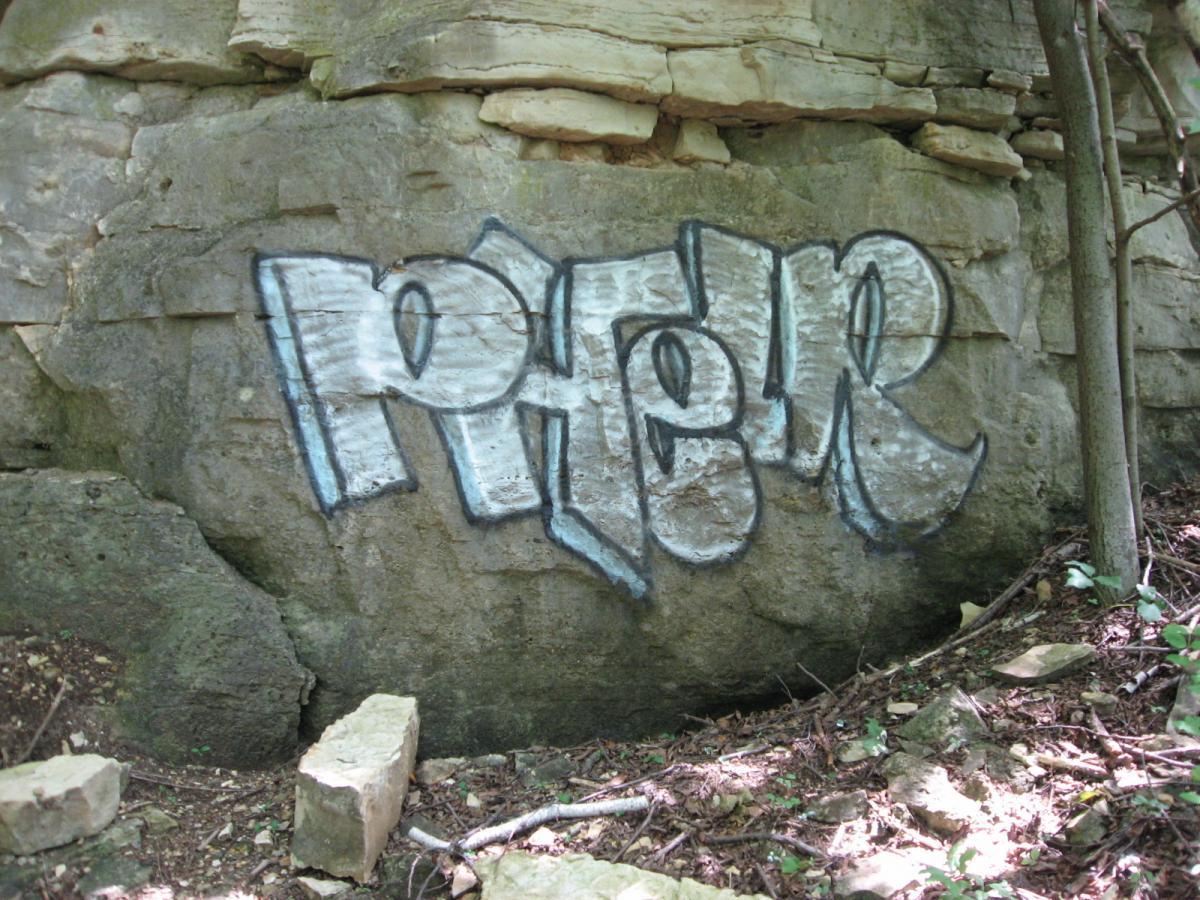 Graffiti artwork on a rock surface in a wooded area, featuring the stylized word "Paper" in white and blue spray paint. Surrounding the rock are small stones and foliage, illustrating a natural setting. Bluff Trails mountain bike trail.