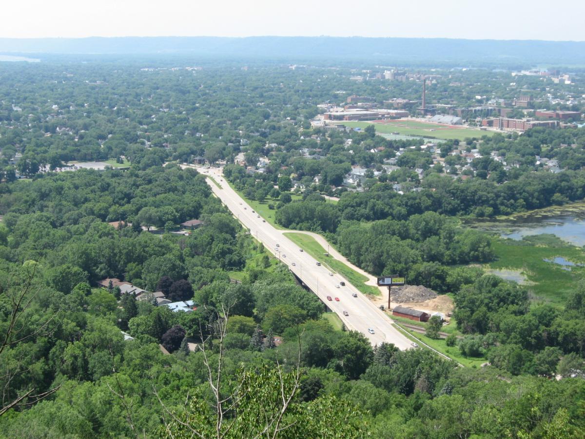 Aerial view of a lush green landscape featuring a highway bordered by trees, with a residential area visible nearby. In the distance, a town with several buildings and a sports field can be seen, along with a body of water. The scene captures the blend of urban and natural environments under a clear sky. Bluff Trails mountain bike trail.