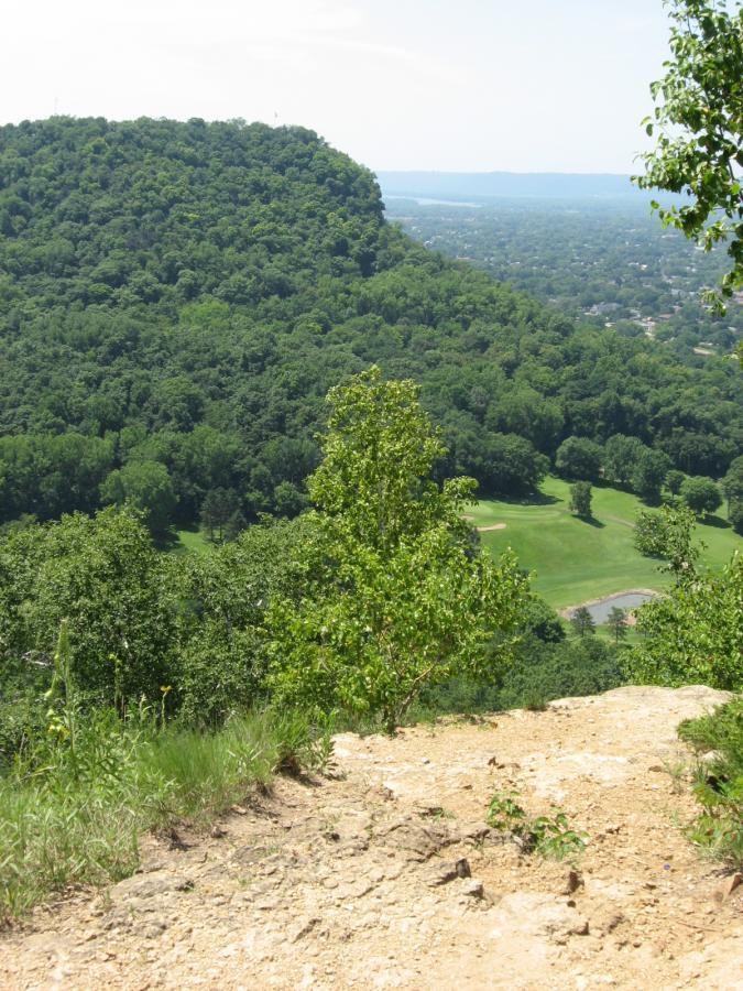 A panoramic view from a rocky hillside overlooking lush green valleys and a distant golf course, with a clear blue sky above. Bluff Trails mountain bike trail.