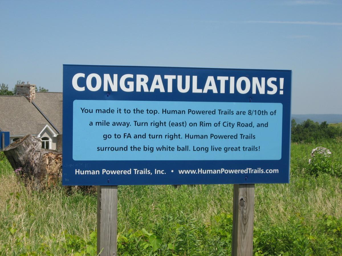 A blue sign displaying the text "CONGRATULATIONS!" at the top, followed by a message indicating directions to the Human Powered Trails, which are 8/10 of a mile away. It instructs to turn right on Rim of City Road and mentions nearby features, also including the website, "www.HumanPoweredTrails.com." The sign is mounted on a wooden post in a grassy area with trees in the background. Bluff Trails mountain bike trail.