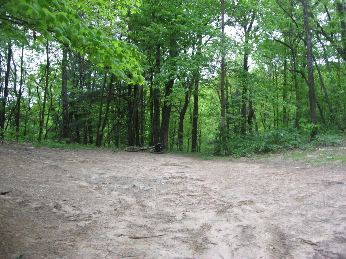 A dirt path forked in a wooded area, surrounded by lush green trees. A bicycle lies on its side near the edge of the path, with tire tracks visible in the soil, indicating recent use. The scene evokes a peaceful and natural outdoor setting. Hickory Nut mountain bike trail.