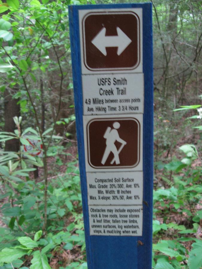 Alt text: A trail sign for USFS Smith Creek Trail, indicating a distance of 4.9 miles between access points and an average hiking time of 3 hours and 45 minutes. The sign lists trail details including compacted soil surface specifications, maximum grade and width, and potential obstacles such as exposed rocks, tree roots, and uneven surfaces. The background features lush green foliage. Hickory Nut mountain bike trail.