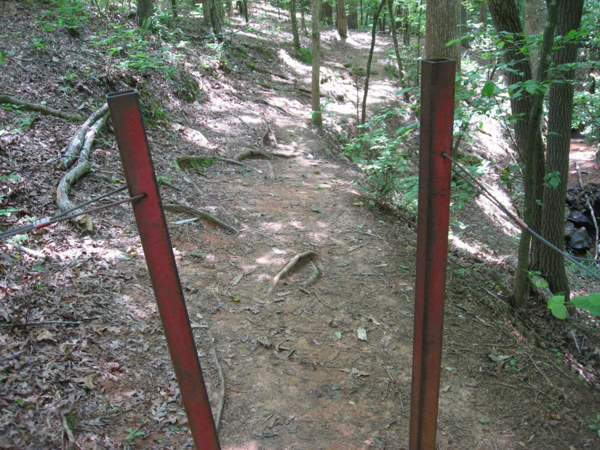 A narrow dirt path through a wooded area, framed by two vertical red metal poles with a wire strung across them. The ground is covered in fallen leaves and tree roots are visible along the sides of the trail. Sunlight filters through the trees, casting dappled shadows on the ground. Lake Herrick mountain bike trail.