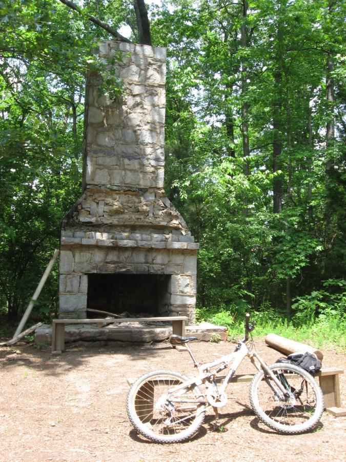 A weathered stone chimney standing amidst lush green trees, with a mountain bike leaning against a wooden bench in the foreground. The scene is filled with natural sunlight filtering through the leaves, highlighting the rustic charm of the chimney and the surrounding forest. Lake Herrick mountain bike trail.