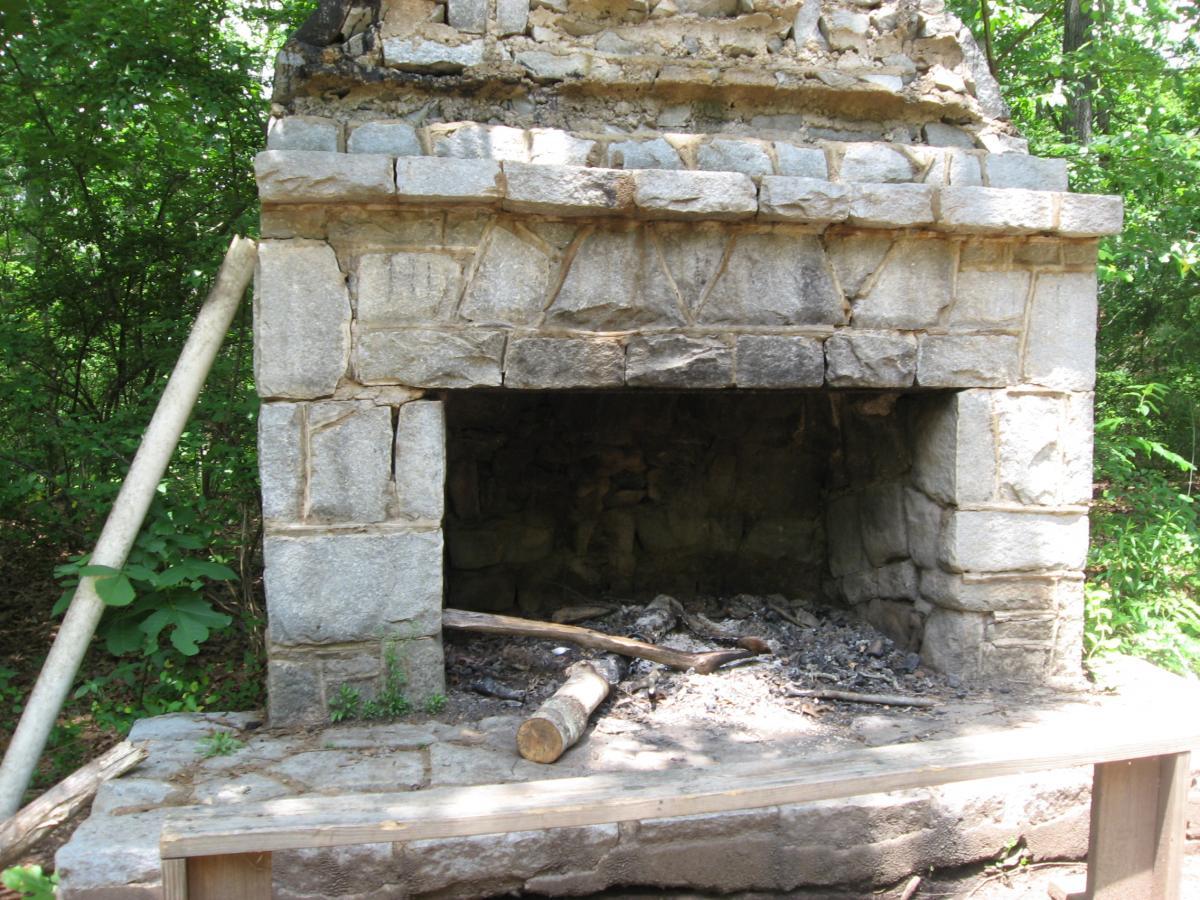 Stone fireplace structure in a wooded area, partially overgrown with vegetation. The fireplace is made of rough stones and has a wide opening, with remnants of ash and logs inside. A wooden support beam is visible to the left, and the surrounding trees are lush and green. Lake Herrick mountain bike trail.