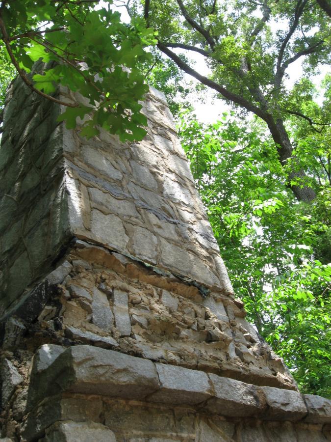 A weathered stone structure partially covered by green foliage, set against a background of trees and bright sunlight. The image captures the texture of the stonework, highlighting its rough, irregular surfaces, and conveys a sense of age and nature. Lake Herrick mountain bike trail.