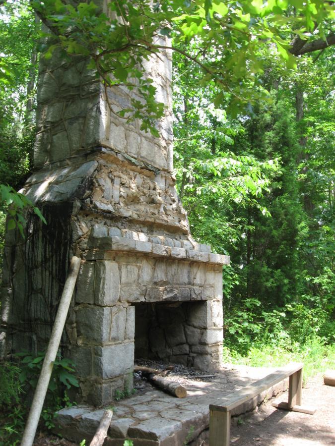An old, weathered stone chimney stands in a lush, green forest. The structure shows signs of deterioration, with exposed rock and missing masonry. Surrounding the chimney are dense trees and foliage, creating a natural setting. A flat stone base supports the chimney, with logs placed nearby, suggesting it may have been part of an old outdoor gathering area. Lake Herrick mountain bike trail.