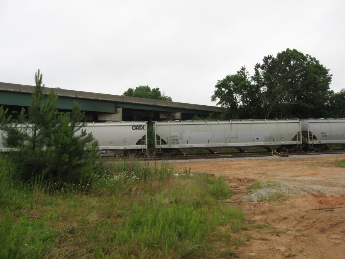 A train with several gray freight cars is positioned near a concrete overpass, surrounded by grassy areas and small trees. The sky is overcast, creating a muted atmosphere. Chase Street mountain bike trail.