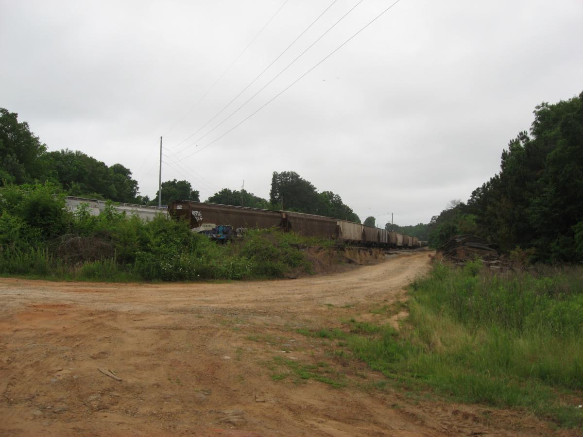 A dirt road leading into a rural area, with overgrown vegetation on either side. In the background, a line of freight train cars can be seen, partially obscured by bushes and trees. The sky is overcast, hinting at a cloudy day. Chase Street mountain bike trail.