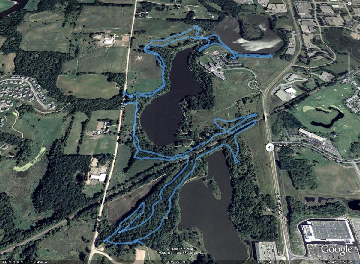 Aerial view of a landscape featuring a winding river and surrounding wooded areas, with marked blue lines indicating trails or pathways. Residential areas and fields are visible in the background, along with nearby roads and commercial buildings. Mammoth Trail mountain bike trail.