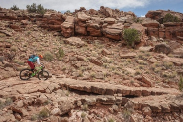 A mountain biker navigating a rocky trail in a desert landscape, surrounded by boulders and sparse vegetation under a partly cloudy sky. Moab Brand Trails mountain bike trail.