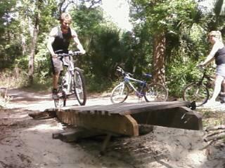 Two cyclists navigate a wooded trail. One rider is jumping over a small wooden ramp, while the other is nearby, positioned to the side with a bicycle. Lush greenery surrounds the path, creating a natural outdoor setting. Chuck Lennon Park mountain bike trail.