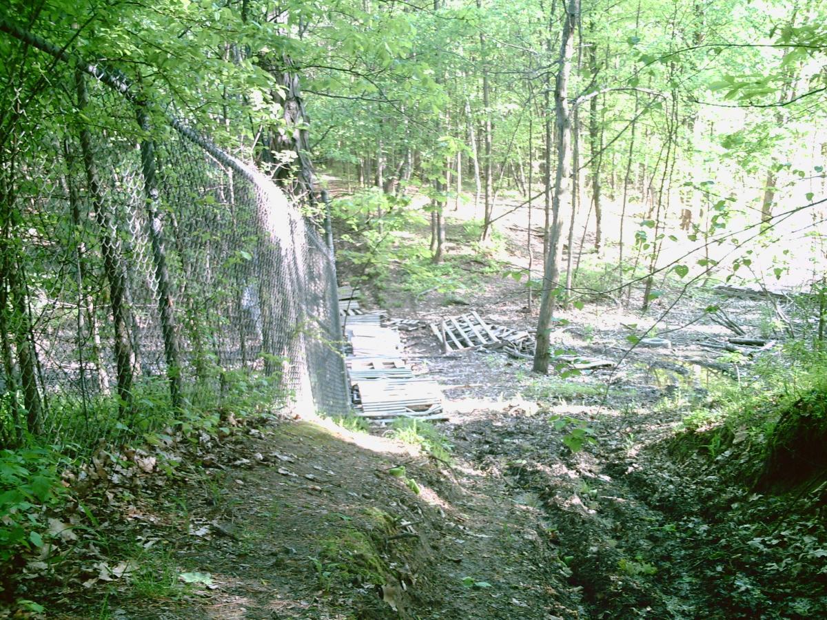 A fenced area next to a wooded trail, with a forested backdrop featuring green foliage and trees. In the foreground, a dirt path lined with fallen leaves leads toward the fence, while wooden pallets and debris are visible in the background. Water Company Owned Woods mountain bike trail.