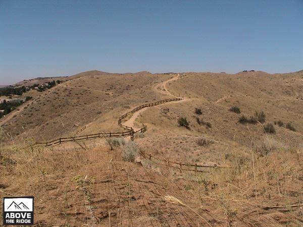 A scenic view of rolling hills and a winding path surrounded by dry grass and sparse vegetation, with a wooden fence along the trail. The clear blue sky above creates a bright, sunny atmosphere. Red Fox Loop mountain bike trail.