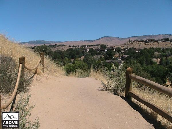 A dirt path lined with wooden rails leads through a hillside with dry grasses and shrubs, providing a view of a residential area and mountains in the background under a clear blue sky. Red Fox Loop mountain bike trail.