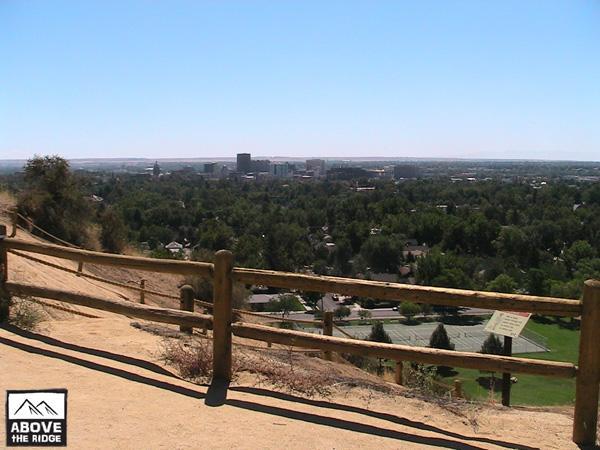 Scenic view from a ridge overlooking a city surrounded by greenery, featuring a dirt path and wooden railing in the foreground, with a clear blue sky above. Red Fox Loop mountain bike trail.