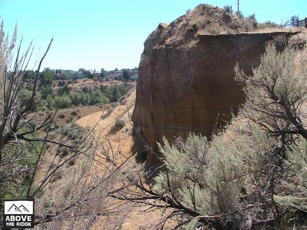 A rocky cliffside rises from a dry landscape, with sparse vegetation and trees in the background. The blue sky above is clear and bright. The scene captures a natural terrain featuring rugged landforms and brush, symbolizing a serene yet rugged outdoor environment. Red Fox Loop mountain bike trail.