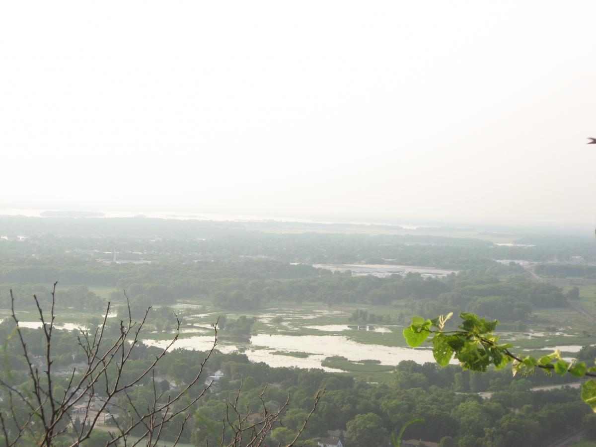 A panoramic view of a misty landscape, showcasing a river winding through green fields and patches of water, with trees and houses dotting the foreground. The sky is overcast, casting a soft, muted light over the scene, enhancing the tranquil atmosphere. Bluff Trails mountain bike trail.