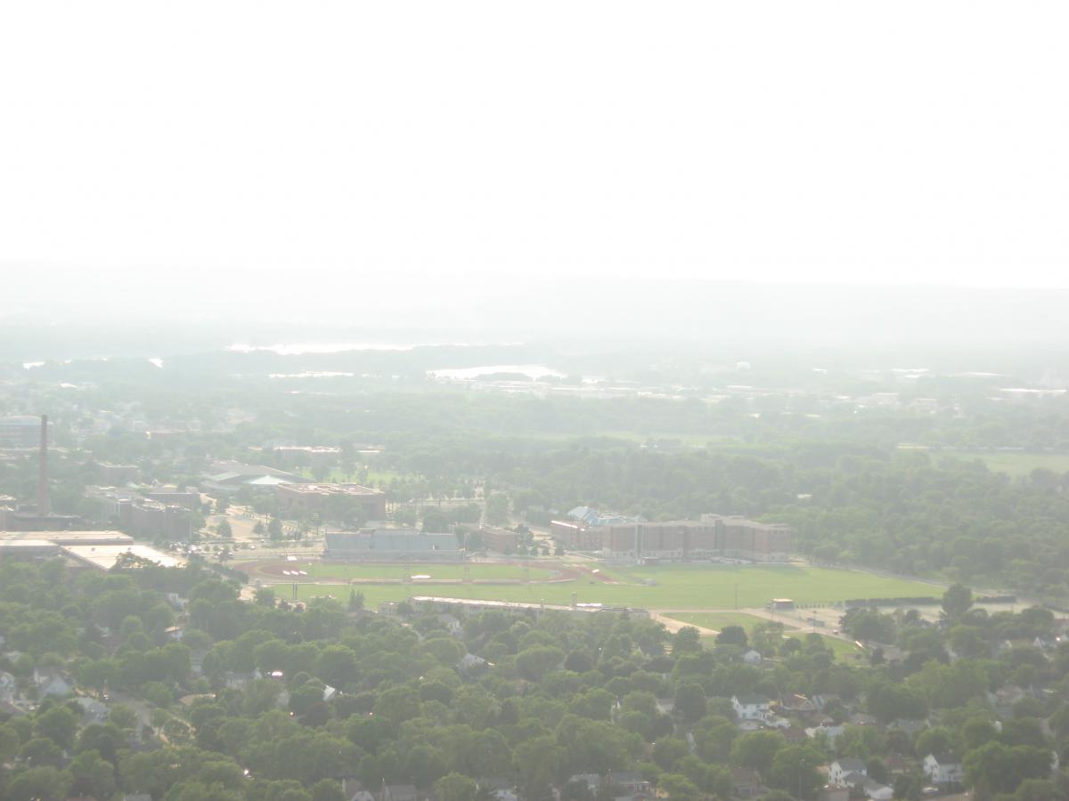 A hazy aerial view of a suburban landscape featuring green trees, a field, and various buildings, with a light mist covering the distant horizon. Bluff Trails mountain bike trail.