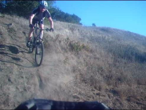 A mountain biker navigating a dusty downhill trail on a sunny day, with a clear blue sky in the background and a grassy hillside. Briones Regional Park mountain bike trail.