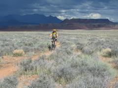 A cyclist riding a mountain bike along a dirt path in a vast, open landscape with sparse vegetation. The backdrop features distant mesas and clouds, creating a dramatic sky. J.E.M. Trail mountain bike trail.