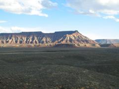 A panoramic view of a rugged mountain landscape featuring layered rock formations under a blue sky with scattered clouds. The foreground consists of flat, sparse terrain that gradually rises to the distinctive mesas in the background. J.E.M. Trail mountain bike trail.