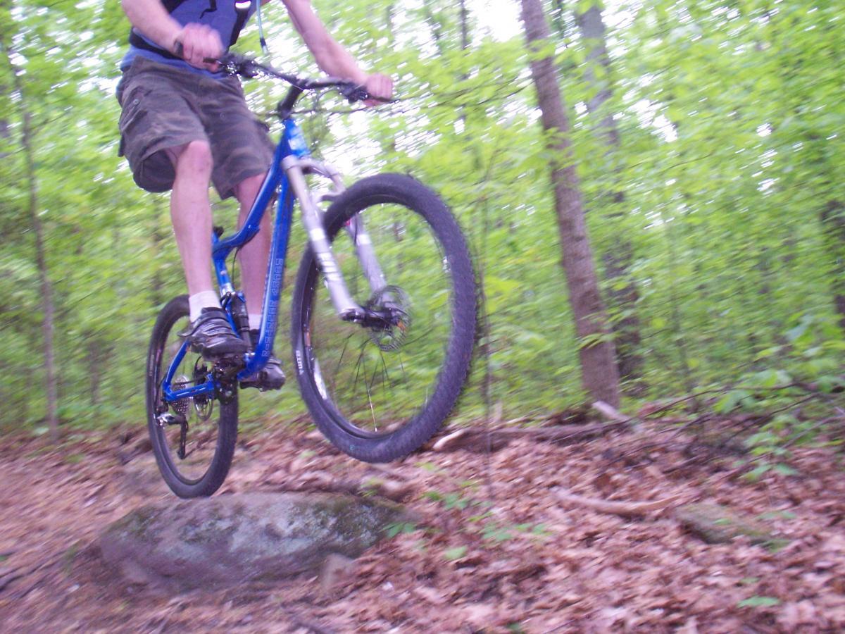 A person is mid-air while riding a blue mountain bike, airborne above a rock on a forest trail. The background features green trees and leaves on the ground, illustrating an outdoor biking scene. Powhite Park mountain bike trail.