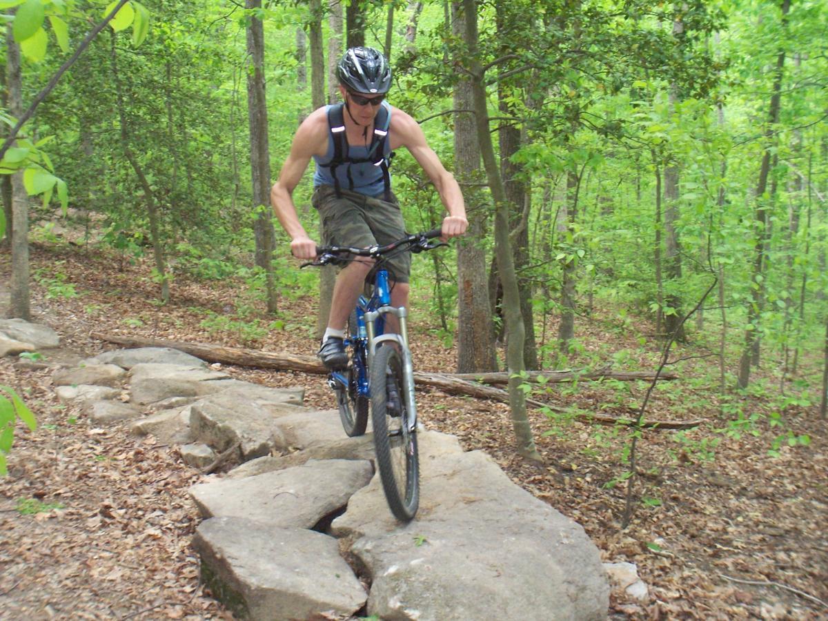 A person riding a mountain bike over rocky terrain in a lush, green forest. The individual is wearing a helmet and is dressed in a tank top and shorts, focused on navigating the path. Surrounding trees and foliage create a vibrant outdoor setting. Powhite Park mountain bike trail.