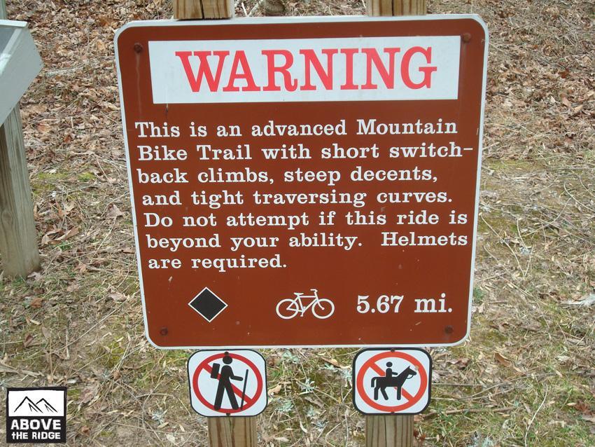 A warning sign for an advanced mountain bike trail, detailing its features such as short switchback climbs, steep descents, and tight traversing curves. The sign advises riders not to attempt the trail if it exceeds their ability and states that helmets are required. Below the main message, there are icons indicating restrictions against pedestrians and horses on the trail, along with the trail length of 5.67 miles. York River State Park mountain bike trail.