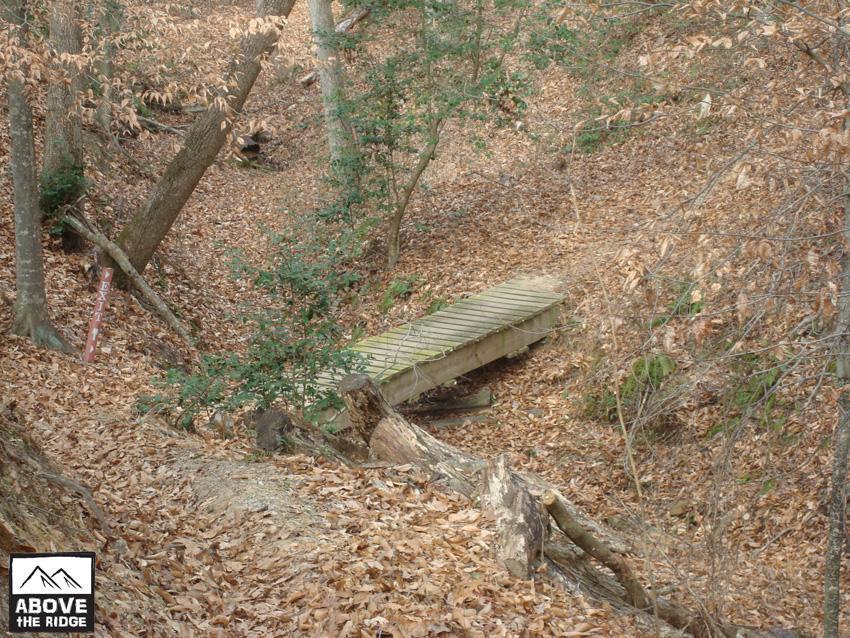 A wooden bridge spans a small ravine, surrounded by trees and scattered autumn leaves. The ground is covered in dry leaves, and a trail marker can be seen in the background. The scene conveys a peaceful, natural setting. York River State Park mountain bike trail.