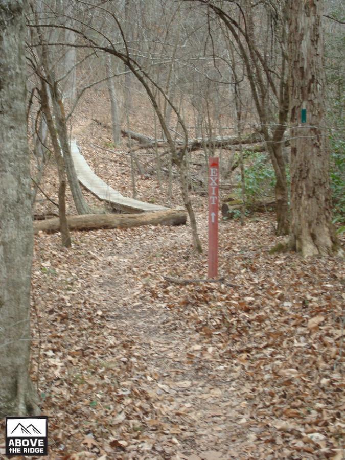 A wooded trail with fallen leaves and trees, featuring a prominent red sign labeled "EXIT" pointing upward. In the background, a wooden pathway can be seen leading through the forest. York River State Park mountain bike trail.