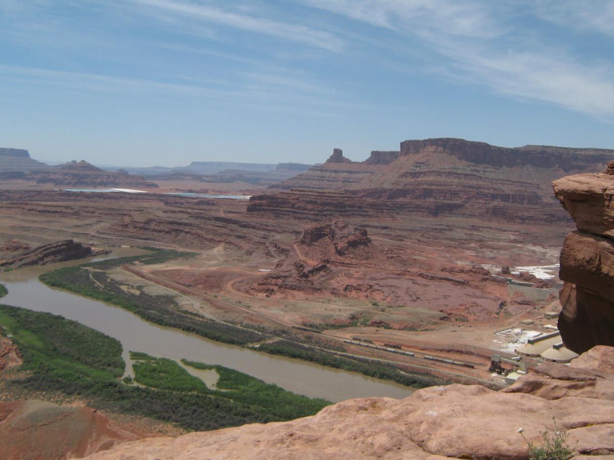 A panoramic view of a rugged desert landscape featuring red rock formations, a winding river, and verdant vegetation along the riverbanks under a clear blue sky. The scene captures the natural beauty of the terrain with distant mesas and cliffs visible in the background. Amasa Back Trail mountain bike trail.