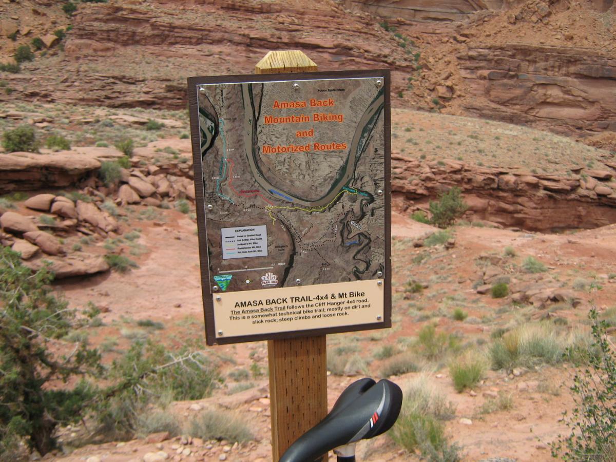 A trail sign for the Amasa Back Mountain Biking and Motorized Routes, featuring a detailed map of the area. The sign includes names of trails, descriptions of their difficulty levels, and information about the terrain. In the foreground, a bicycle seat is partially visible, with red rock formations and sparse vegetation in the background. Amasa Back Trail mountain bike trail.