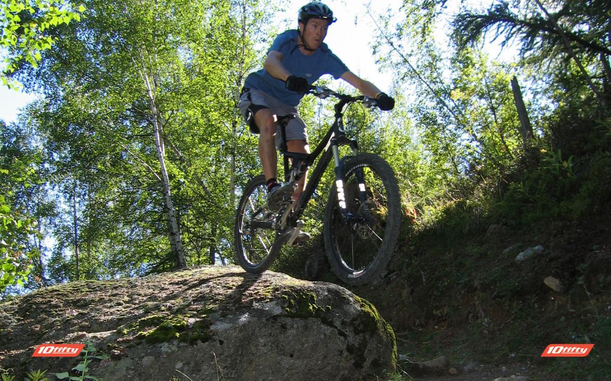 A mountain biker jumping off a large rock on a forest trail, with green trees and foliage in the background. The rider is wearing a helmet and athletic gear, showcasing an action-packed moment in outdoor biking.