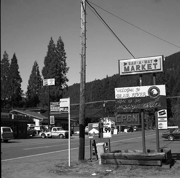 Black and white photograph of a roadside market sign that reads "Sav-A-Way Market" and "Welcome to Blue River, Home of the McKenzie Eagles." The scene includes parked vehicles, a utility pole, and surrounding tall trees, with a highway visible in the foreground. The sign indicates that the market is open.