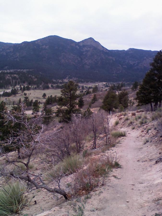 A dirt path winding through a rugged landscape, flanked by sparse vegetation and dry bushes. In the background, a range of mountains rises under a cloudy sky, creating a serene outdoor scene. Falcon Trail mountain bike trail.